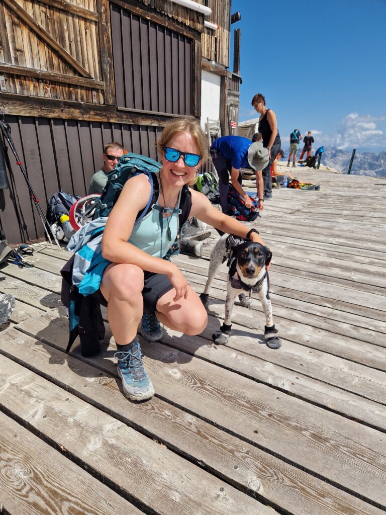 Sportiška mergina atsitūpusi ir apsikabinusi šuniuką Via ferrata Ivano Dibona maršrute
