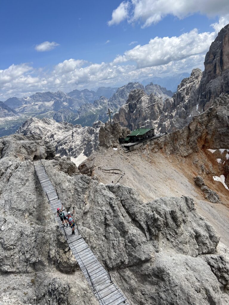 Trobelė kalnuose ir šalia jos kabantis Ponte Cristallo tiltas su dviem merginomis