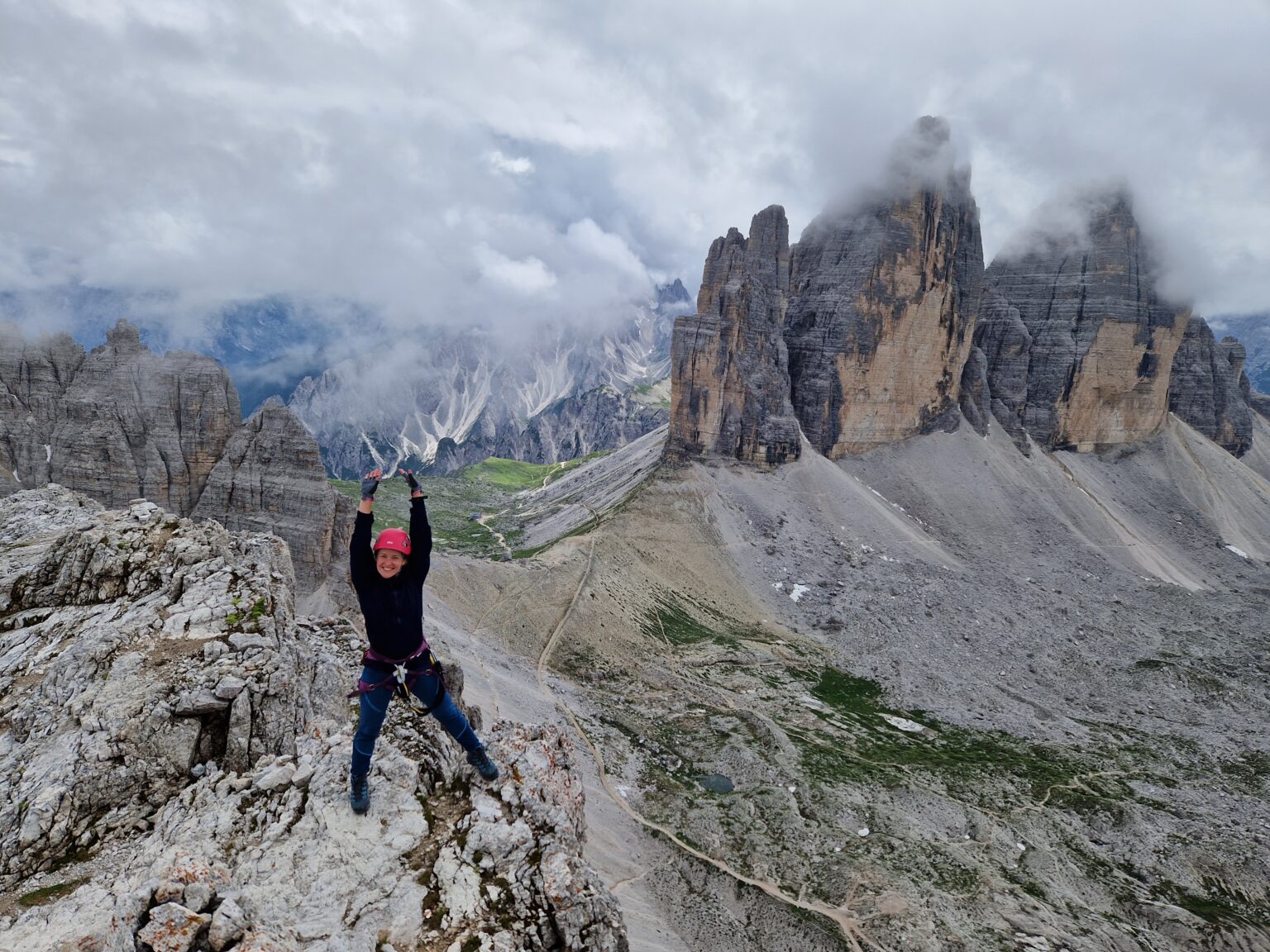 Mergina su rožiniu šalmu stovi ant kalno iškėlusi rankas su Tre Cime di Lavaredo bokštais fone