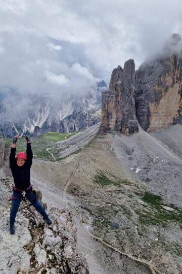 Mergina su rožiniu šalmu stovi ant kalno iškėlusi rankas su Tre Cime di Lavaredo bokštais fone