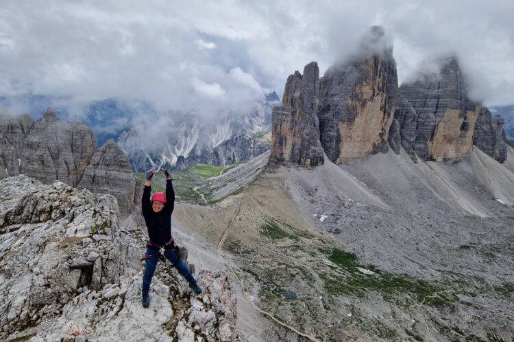 Mergina su rožiniu šalmu stovi ant kalno iškėlusi rankas su Tre Cime di Lavaredo bokštais fone