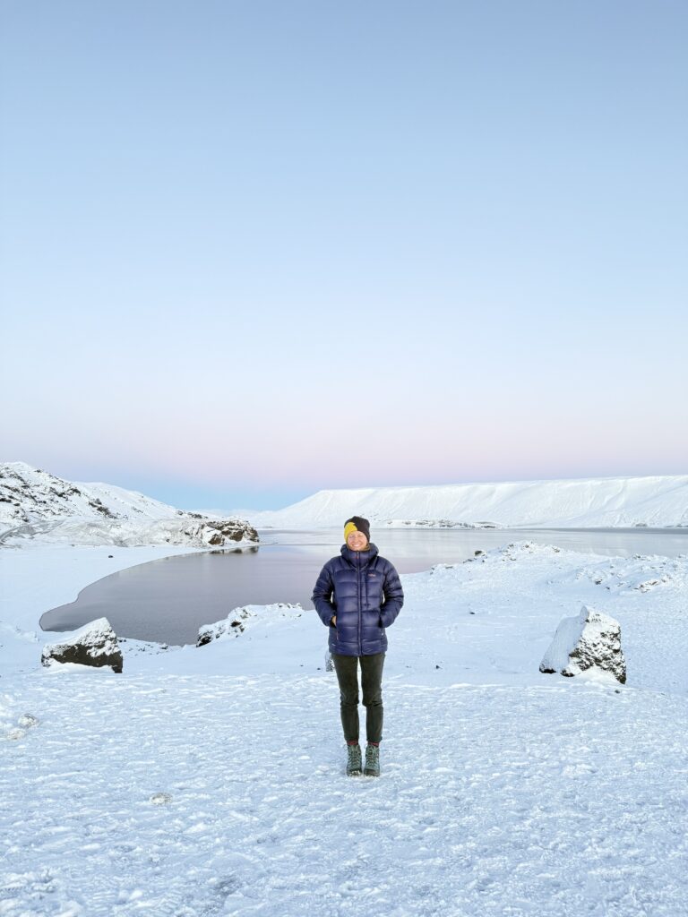 Kelionė į Islandiją žiemą. Moteris su pūkine striuke stovinti ant snieguoto šlaito Islandijoje su rožiniu saulėlydžio dangumi fone