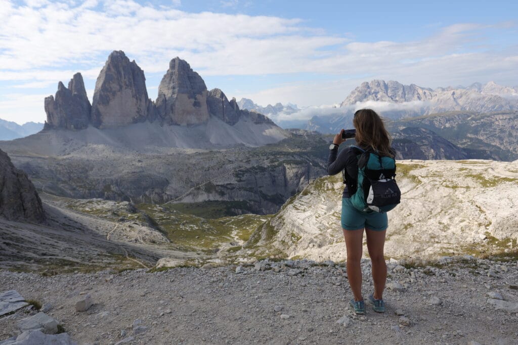 A woman in blue shorts and Black Diamond backpack taking picture of the Tre Cime di Lavaredo towers